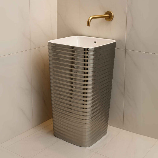 Modern silver pedestal wash basin with ribbed design and square white top, placed in a marble bathroom with gold wall-mounted tap.