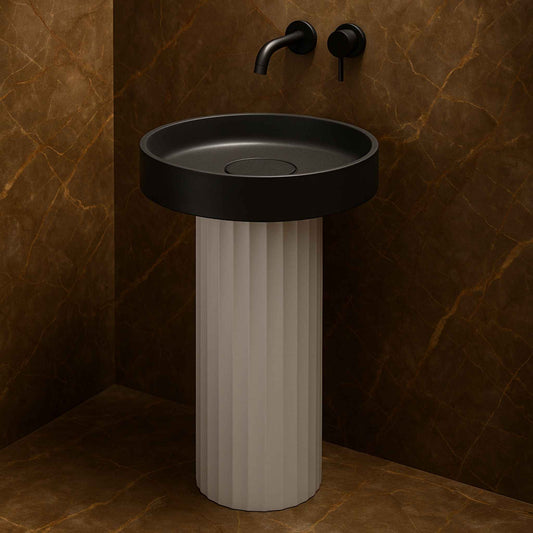  Front view of a round matte black pedestal washbasin with a fluted white base and wall-mounted black faucet in a luxurious marble bathroom.